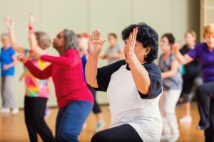 A large group of people learn to Zumba or line dance at senior center. The active seniors clap and dance as they enjoy dancing. The group consists of men and women.